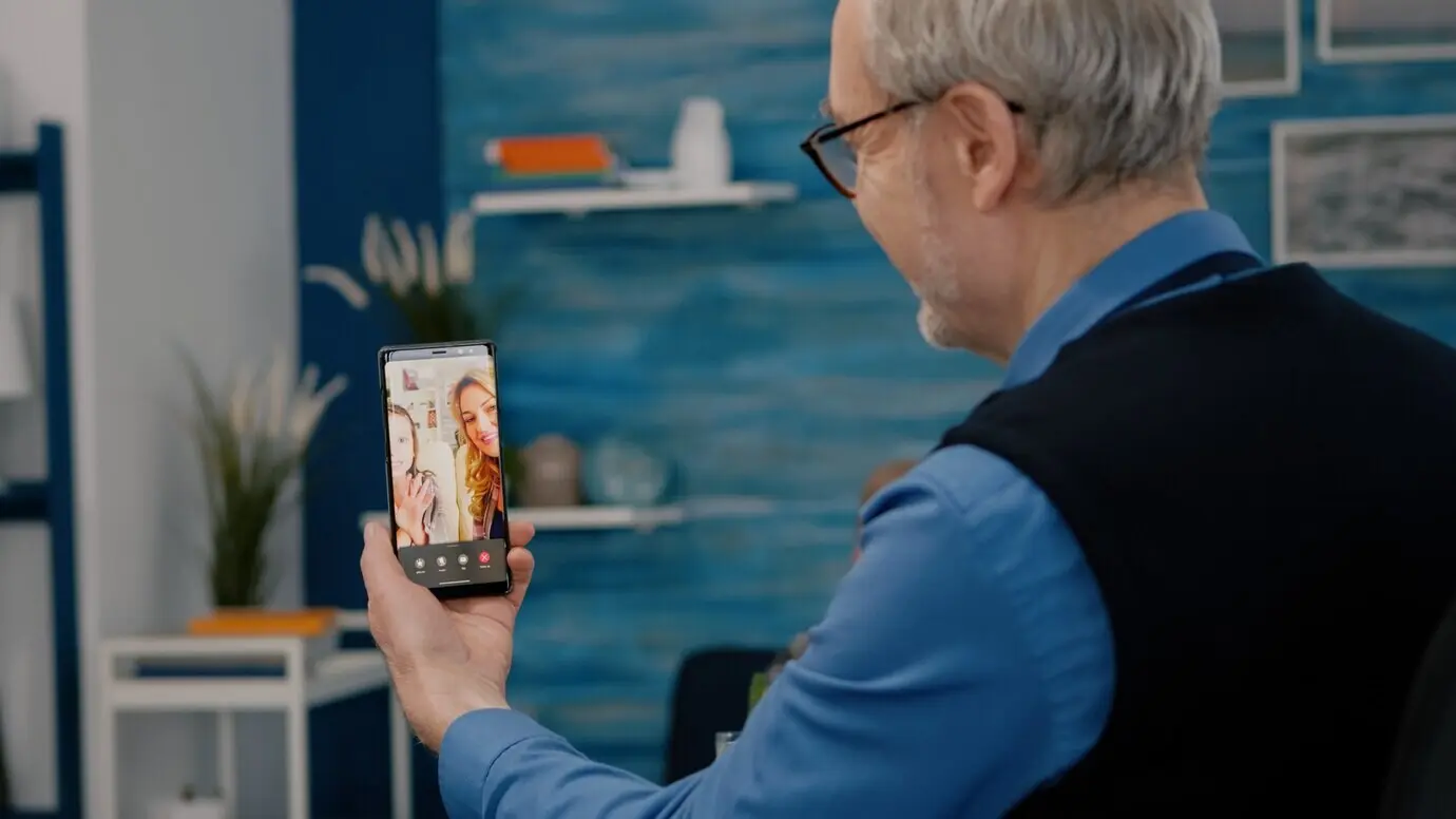 Пожилой мужчина на пенсии сидит в гостиной и с помощью смартфона ведёт онлайн‑видеозвонок с племянником.